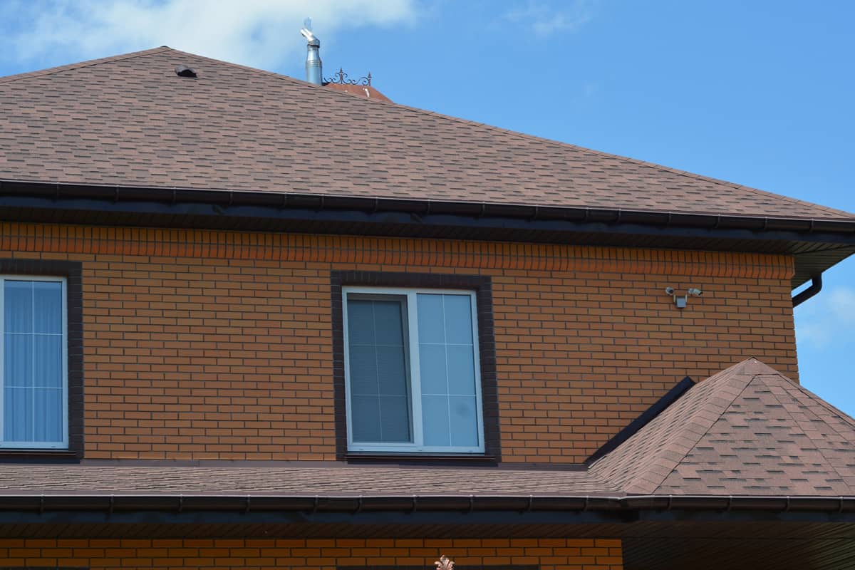 A close-up of a house with an asphalt shingled roofing construction, a roof ventilation cap, plastic soffit, rain gutters, and security cameras installed.