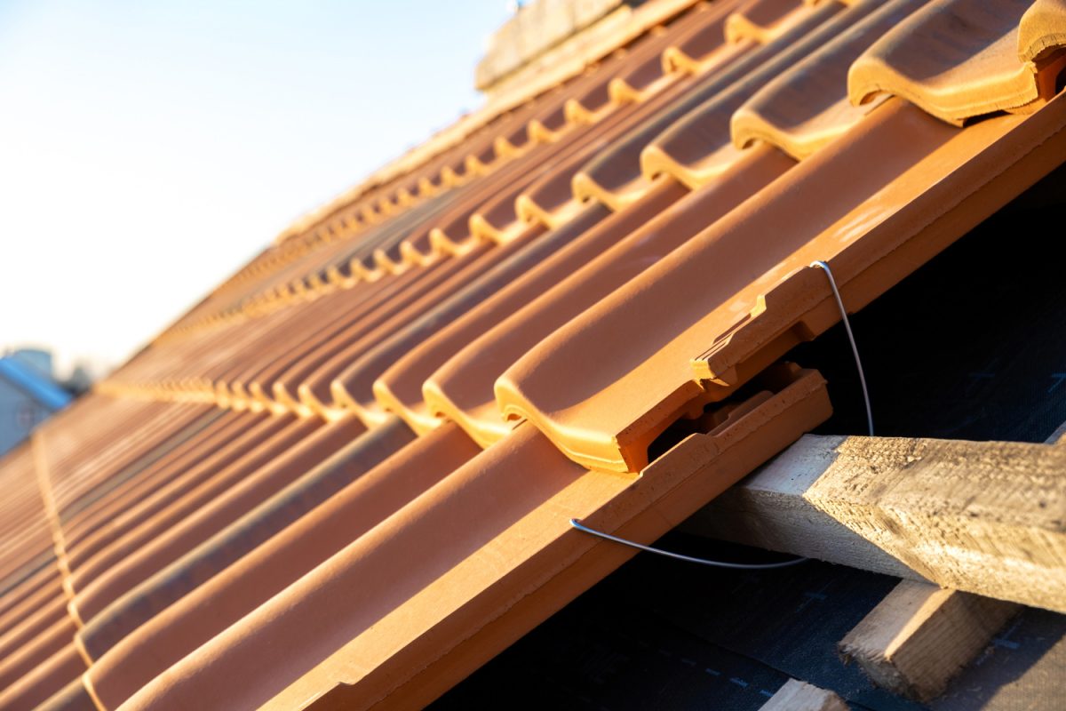 Closeup of metal montage anchor for installation of yellow ceramic roofing tiles mounted on wooden boards covering residential building roof under construction.


