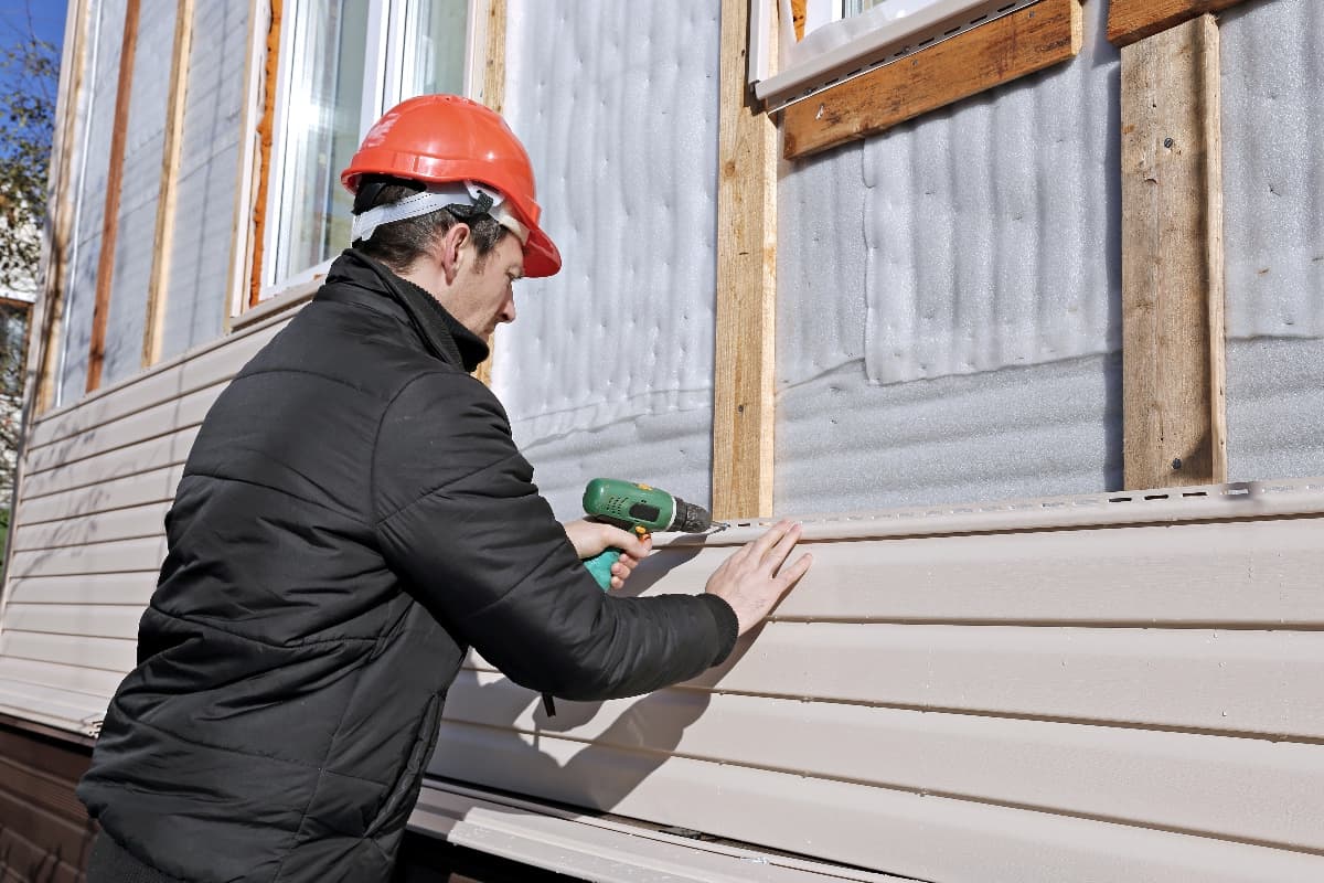 What Are Some Sheathing Materials You Can Use For Vinyl Siding - A worker installs panels beige siding on the facade of the house