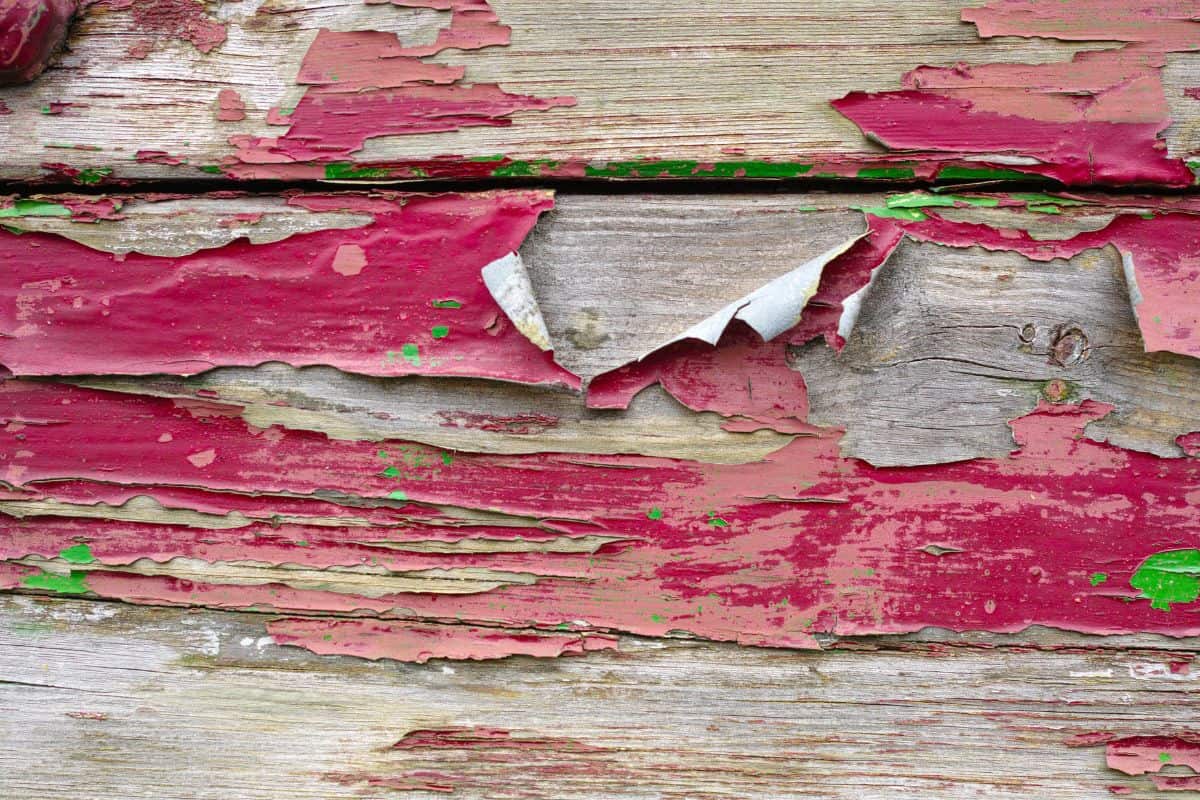 a very old red paint layer on textured wooden planks, some other green paint is visible. The paint is full of cracks and flakes.