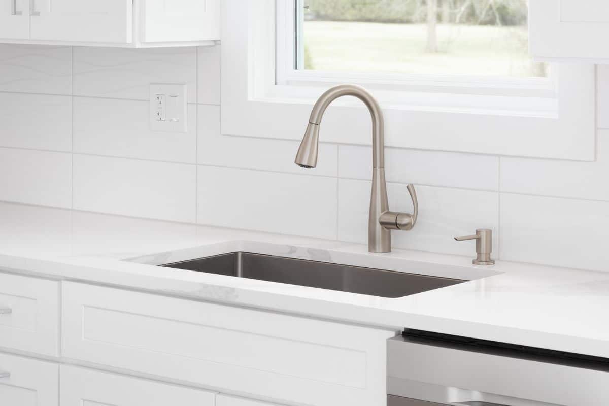 A kitchen sink detail with white cabinets, pendant lights hanging from the ceiling, and a tiled backsplash.
