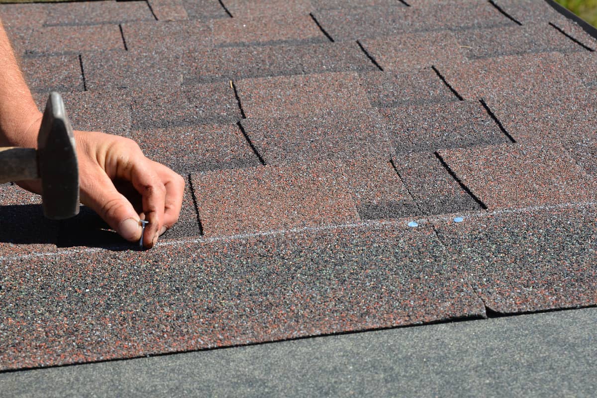 Roofer hands installing asphalt shingles on house construction roof corner with hammer