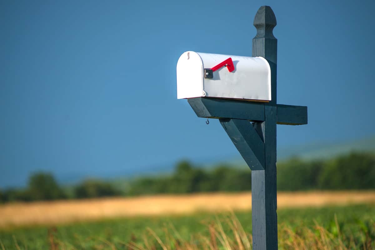 Rural white mailbox with red flag on blue post astride road with field and blue sky in background..